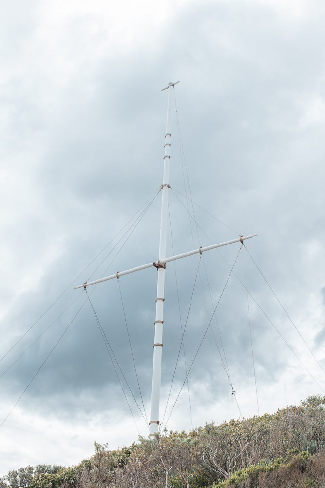 A tall white mast with a horizontal yardarm and rigging stands against a cloudy sky. The mast is secured by multiple guy wires, and it rises from a hill covered in dry brush and green foliage.