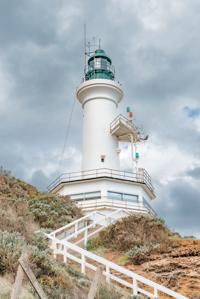 A white lighthouse stands tall on a hill overlooking the ocean, with a cloudy sky above. A white staircase leads up to the lighthouse, surrounded by dry brush and rocks. The lighthouse is equipped with navigational lights and antennas, suggesting its purpose is to guide ships at sea.