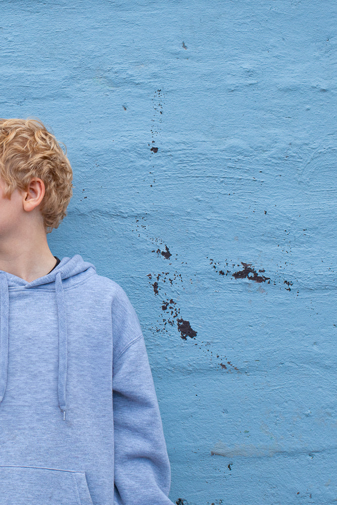 A young boy with curly blonde hair, wearing a light blue hoodie, stands in front of a textured blue wall. The boy is shown from the shoulders up, with his head turned to the left, revealing his ear and the side of his face.