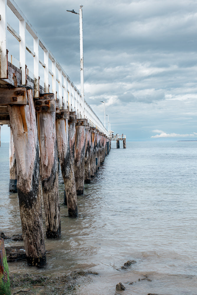 A weathered wooden pier extends into calm water under a cloudy sky. The pier's thick, barnacle-covered pilings are visible, supporting a white railing. A few people are visible at the far end of the pier.