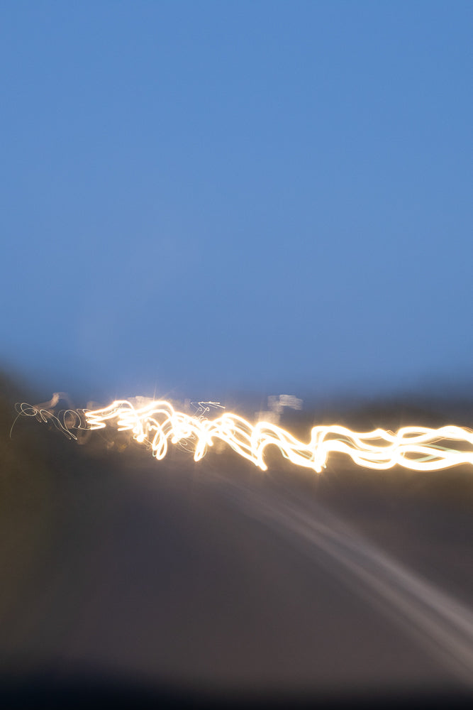 Abstract image of light trails against a blue sky. The bright yellow-white light forms a wavy, serpentine pattern across the lower half of the frame, suggesting movement and speed.