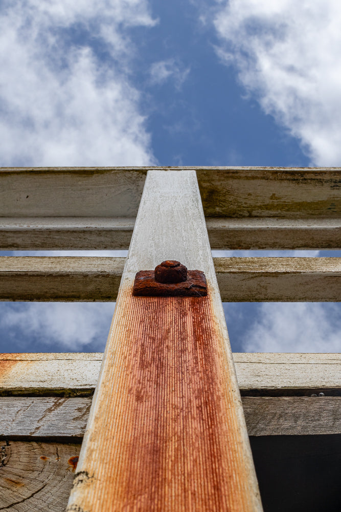 A low-angle shot looking up at a weathered wooden structure against a blue sky with white clouds. A rusty bolt and washer secure a vertical wooden post to horizontal planks.