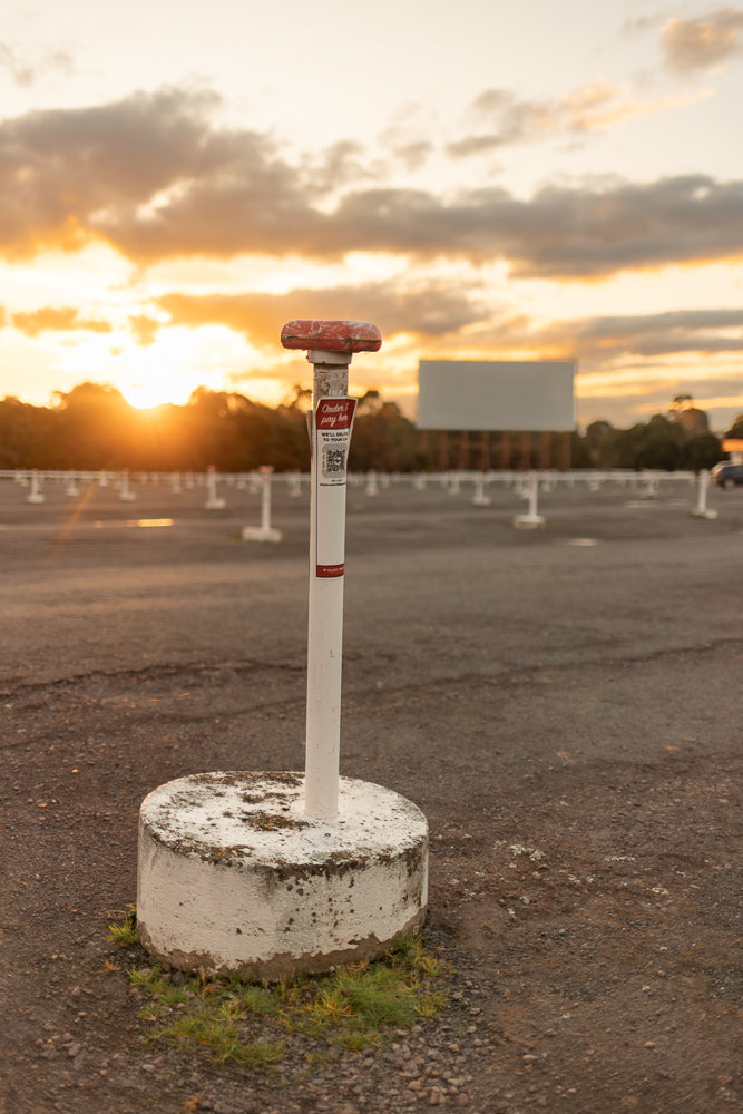 A white post with a red top stands in the foreground of a drive-in movie theater parking lot at sunset. The sky is filled with dramatic clouds illuminated by the setting sun.