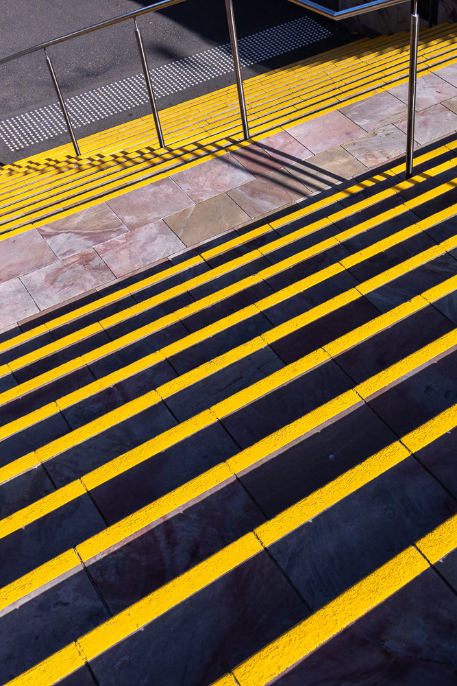 A high-angle view of outdoor stairs with bright yellow safety strips on each step. The sun casts strong shadows across the steps, creating a graphic pattern. A metal handrail runs along the top left, and tactile paving is visible on the landing.