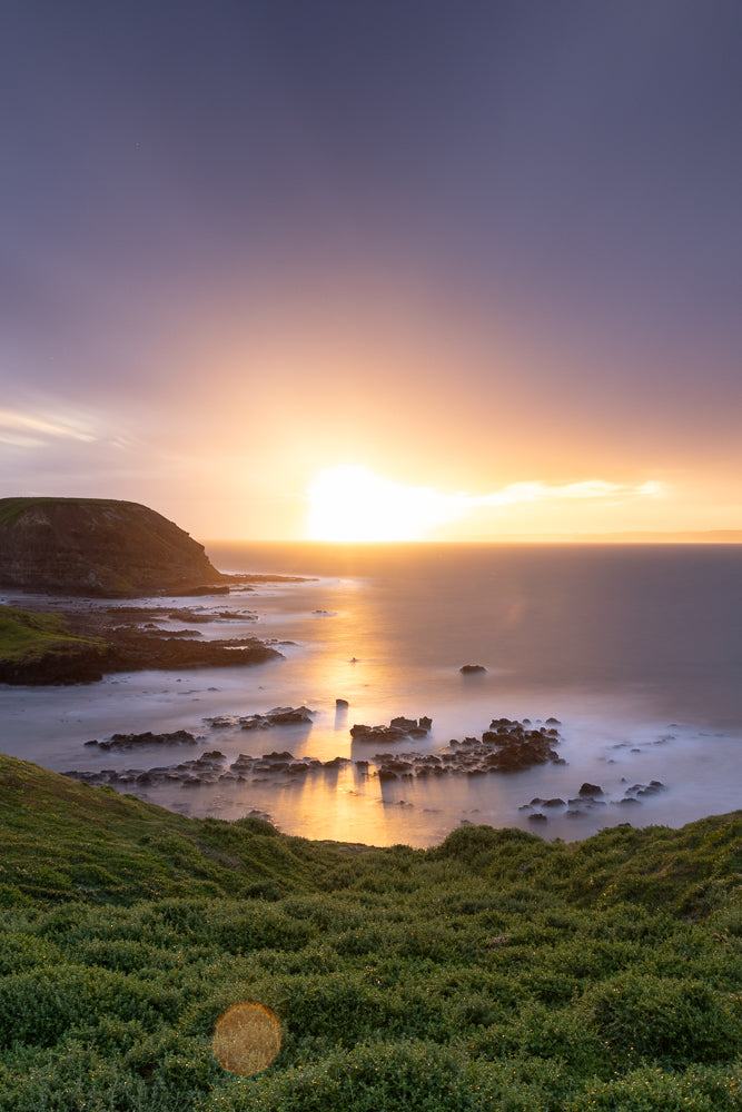 A serene coastal landscape at sunset. The sun, a bright orb, casts a golden glow across the calm ocean, reflecting on the water's surface. Jagged rocks emerge from the misty sea, creating a dramatic contrast with the smooth, blurred water. Lush green grass covers the foreground hills, leading the eye towards the tranquil scene.