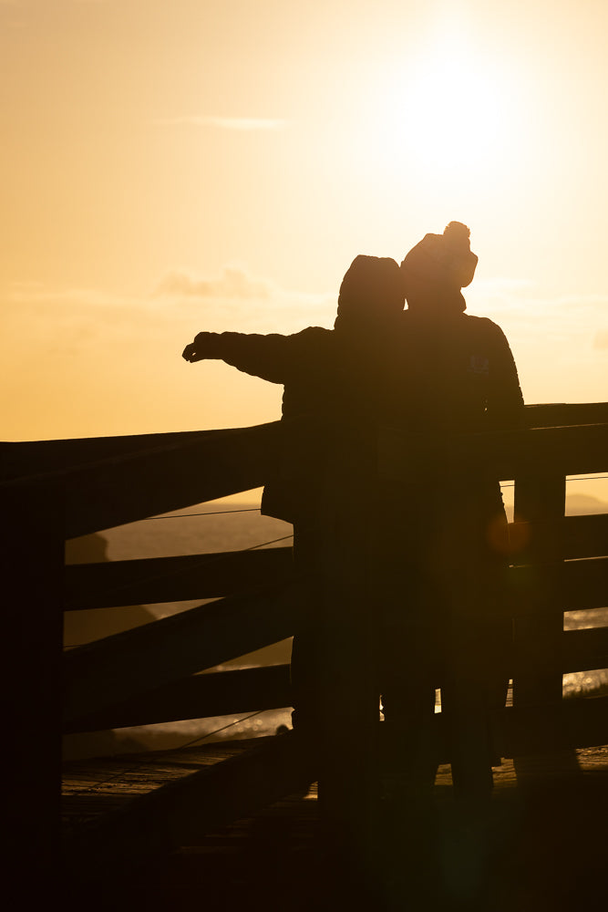 Two people silhouetted against a bright orange sunset, standing on a wooden walkway. One person has their arm outstretched towards the horizon.