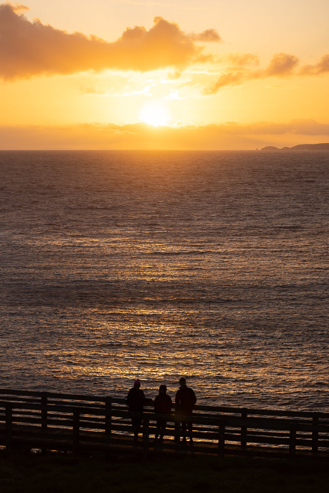 Three people stand on a wooden railing overlooking the ocean at sunset. The sun is setting on the horizon, casting a warm orange glow across the sky and reflecting on the water.