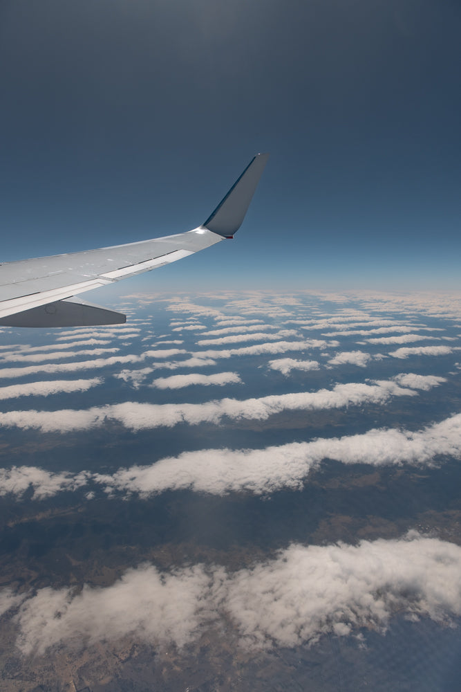 View from an airplane window showing the wing and a layer of fluffy white clouds stretching across the landscape below under a clear blue sky.