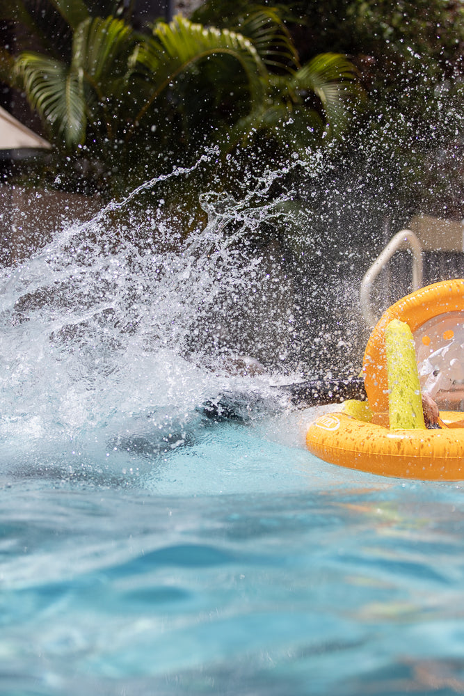A person splashes into a swimming pool, creating a large spray of water. An orange inflatable toy boat with a yellow sail is visible to the right.