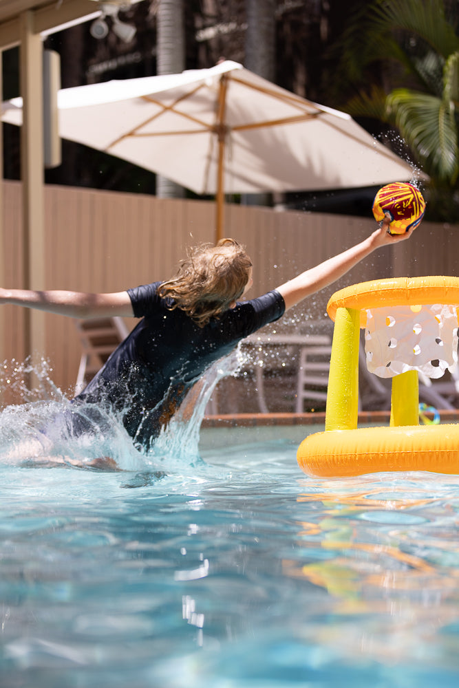 A young person with long blonde hair, wearing a dark t-shirt, splashes into a swimming pool while reaching for a colorful ball to throw into an inflatable basketball hoop.