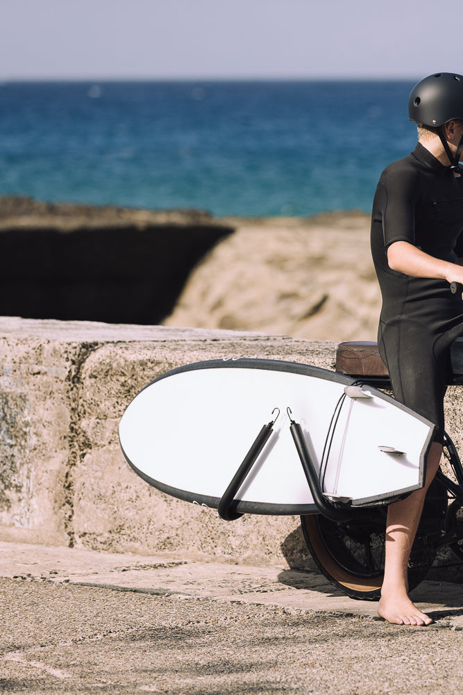 A person in a wetsuit and helmet sits on a bicycle with a surfboard attached to the side. The ocean is visible in the background.