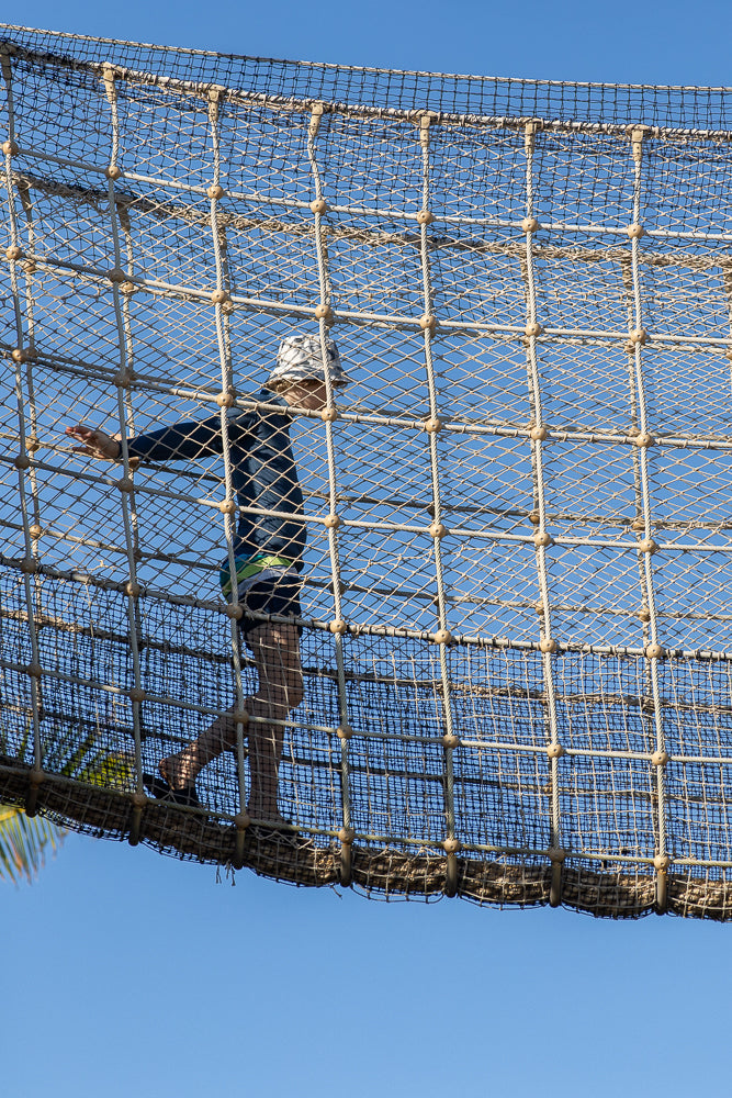 A child wearing a bucket hat and dark blue long-sleeved shirt walks across a rope bridge against a clear blue sky.