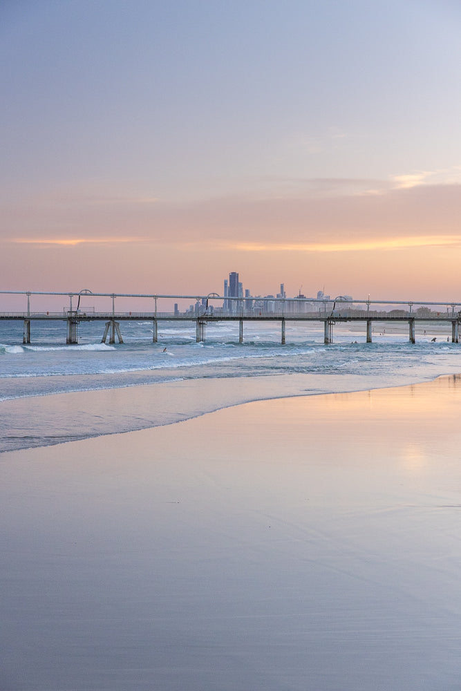 A wide shot of a beach at sunset with a pier extending into the ocean. The sky is filled with soft pink and purple hues, reflecting on the wet sand. Gentle waves roll onto the shore, and a few surfers can be seen in the water.
