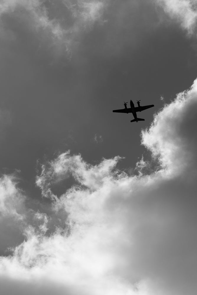 A twin-engine propeller airplane flies through a cloudy sky. The plane is silhouetted against the bright clouds, with its propellers visible.