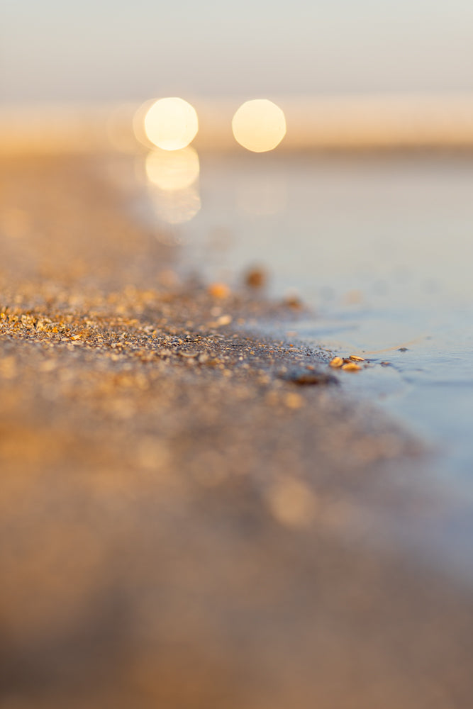 A close-up, low-angle shot of a sandy beach with small pebbles and shells at the edge of the water. The background is blurred with soft, golden bokeh lights.