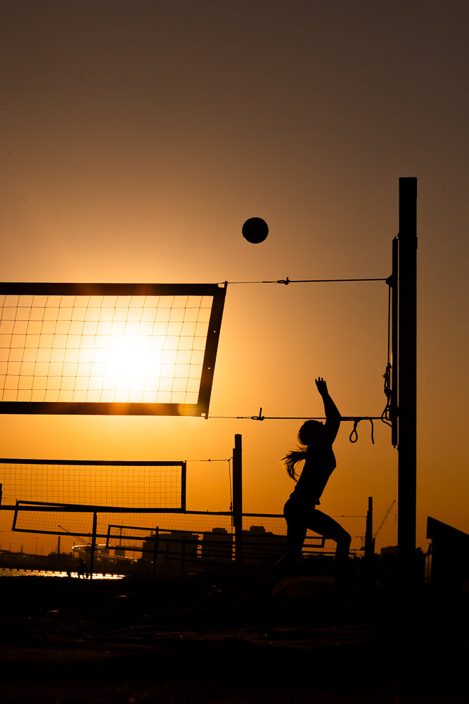Silhouette of a female volleyball player jumping to spike the ball during a sunset game on a beach court.