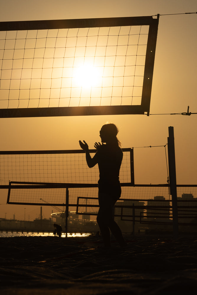 Silhouette of a woman playing beach volleyball at sunset. The sun is low on the horizon, casting a warm glow and creating a dramatic backlight effect.
