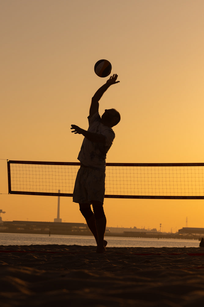 Silhouette of a man playing beach volleyball at sunset. He is in the process of serving the ball, with his arm extended upwards and the ball in the air above him. A volleyball net is visible in the background, along with a hazy shoreline and sky.