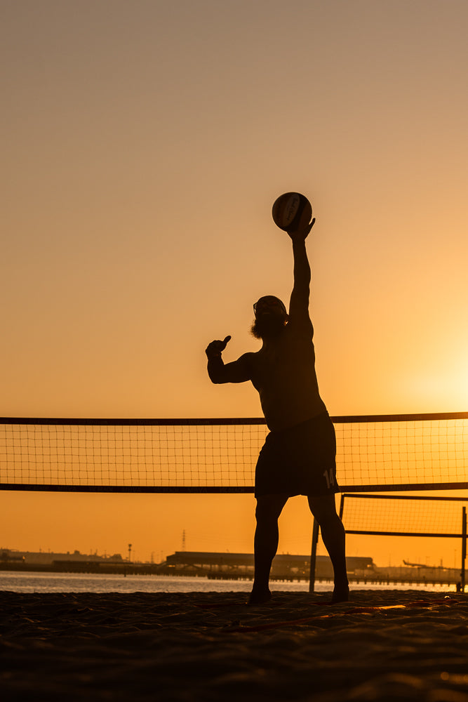 Silhouette of a man serving a volleyball on a beach at sunset. The volleyball net is visible in the foreground, and the sky is a warm orange hue.