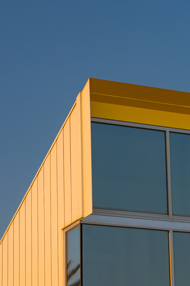 A modern building with a yellow overhang and large windows reflects the blue sky. The building's facade is made of light-colored vertical panels, catching the warm glow of the setting sun.