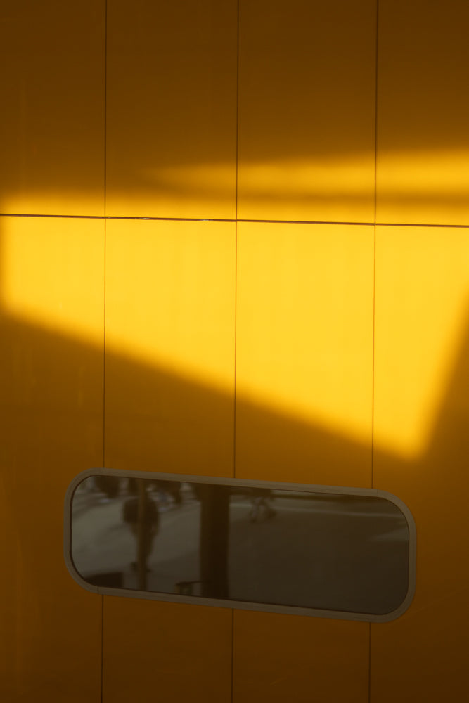 A bright yellow wall with a horizontal band of shadow across the top half. A dark, horizontal, oval-shaped window is centered in the lower half of the wall.