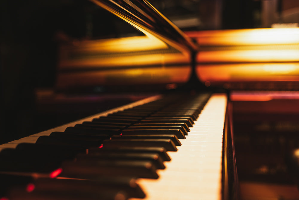 A close-up, low-angle shot of a grand piano's keyboard. The black and white keys are in sharp focus in the foreground, leading the eye towards the blurred, warm, golden-orange background, which reflects light.