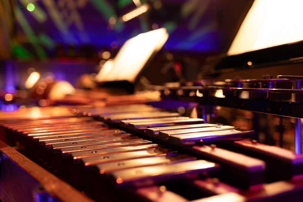 A close-up, low-angle shot of a xylophone with its wooden frame and metal bars illuminated by colorful stage lights. The background is blurred with bokeh effects from purple and green lights.
