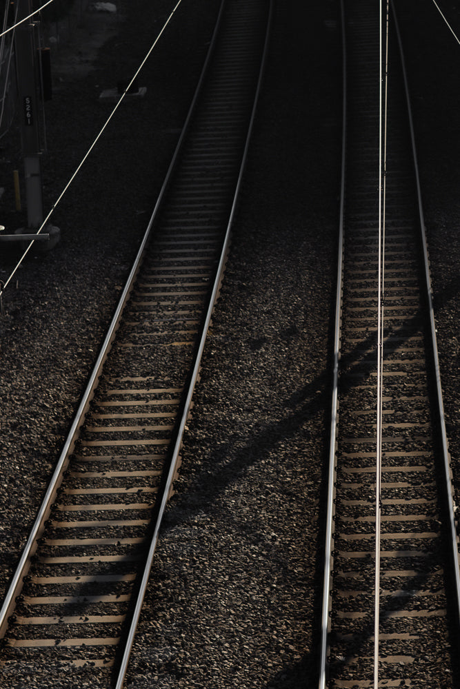 A high-angle shot shows two sets of train tracks receding into the distance. The tracks are made of dark gravel and wooden sleepers, with metal rails. Overhead wires run parallel to the tracks, casting shadows.