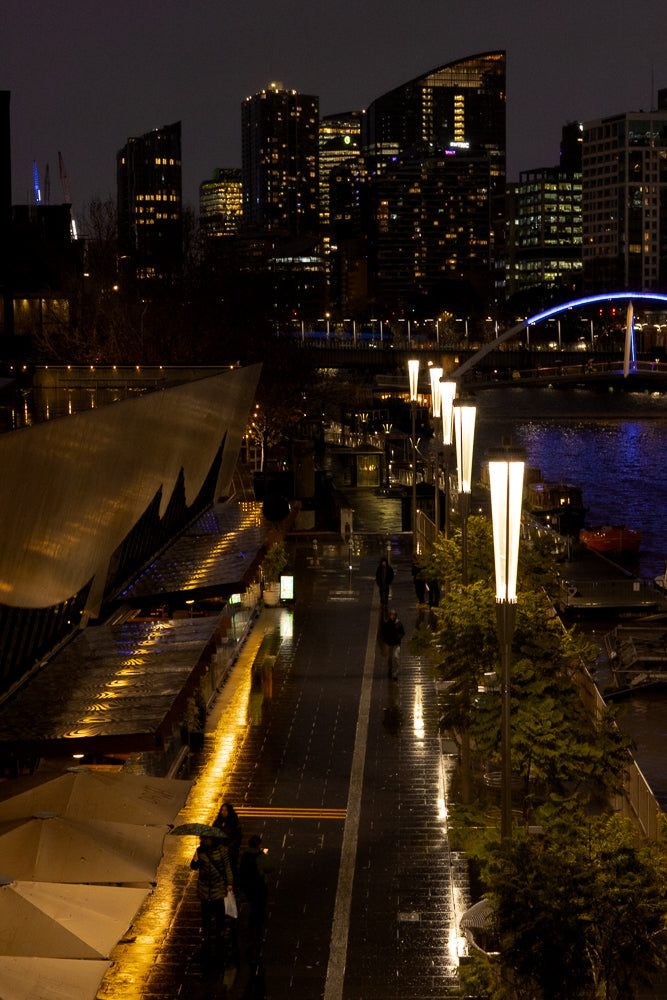 A nighttime cityscape with a waterfront promenade. Tall buildings with illuminated windows dominate the background, while modern streetlights line the wet walkway. Several people are seen strolling along the path, some with umbrellas, reflecting the city lights.