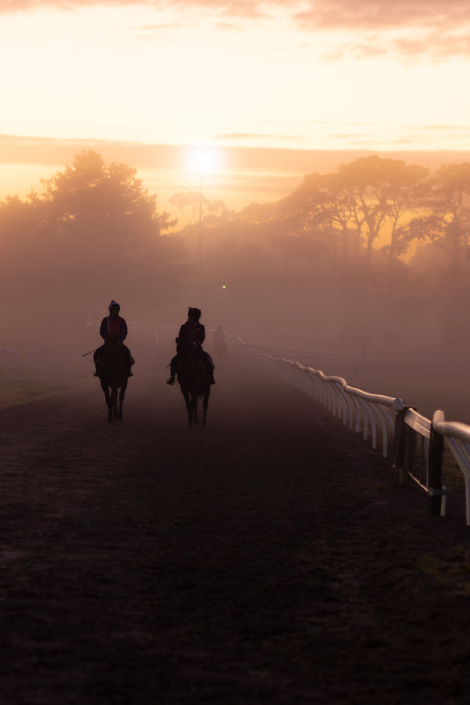Two jockeys on horseback ride along a dirt track at sunrise, shrouded in mist. The sun is low on the horizon, casting a warm glow through the fog and silhouetting the trees in the background.