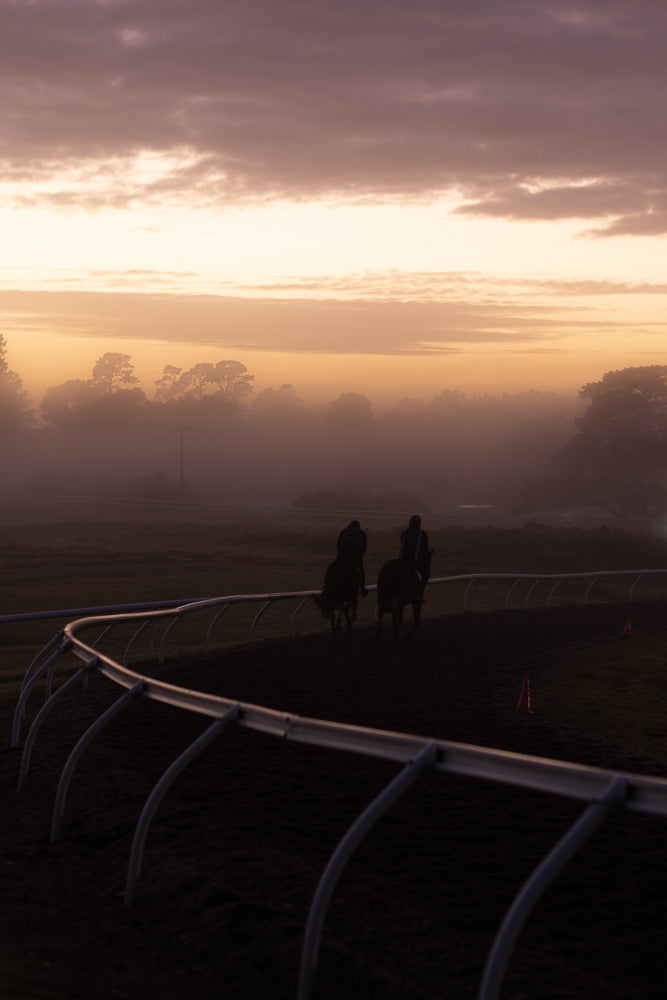 Two jockeys on horseback ride along a track at sunrise. The sky is filled with soft, hazy clouds and the sun casts a warm, golden glow over the misty landscape.