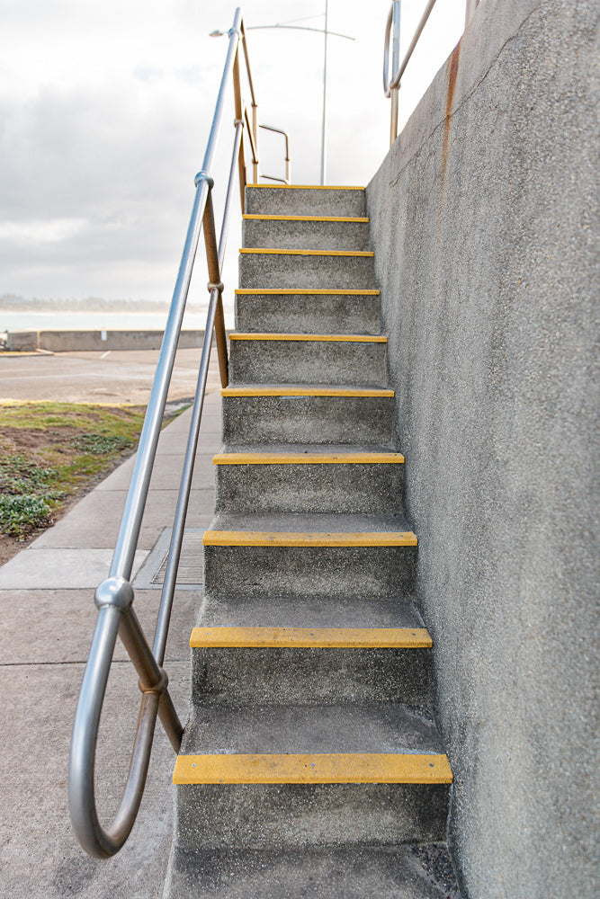 A concrete staircase with yellow anti-slip treads ascends alongside a textured concrete wall. A metal handrail with a curved section is visible on the left. The background shows a paved area, grass, and a body of water under a cloudy sky.