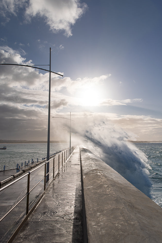 A large wave crashes over a pier walkway on a sunny day. The sun is bright in the sky, partially obscured by clouds. The walkway is wet and reflects the light. A railing runs along the left side of the walkway.