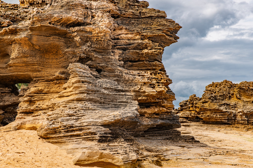 A close-up view of layered, eroded sandstone rock formations on a sandy beach under a cloudy sky. The rocks have a rough, textured surface with many holes and crevices.