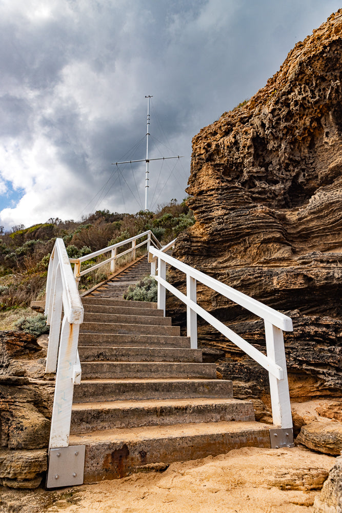 A set of concrete stairs with white railings leads up a sandy embankment towards a tall mast against a cloudy sky. The stairs are weathered and show signs of age, with sand accumulating on the steps. The mast, possibly a flagpole or part of a navigational aid, stands prominently against the dramatic sky, suggesting a coastal location.
