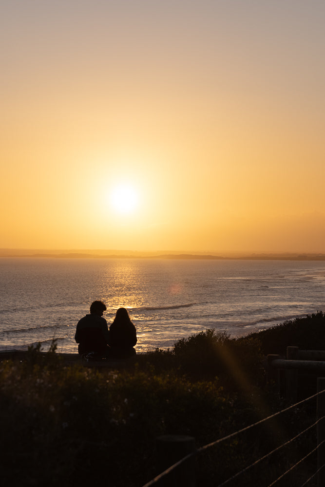 A couple sits on a cliff overlooking the ocean at sunset. The sun is low on the horizon, casting a warm golden glow across the sky and water. Waves gently roll onto the shore below.
