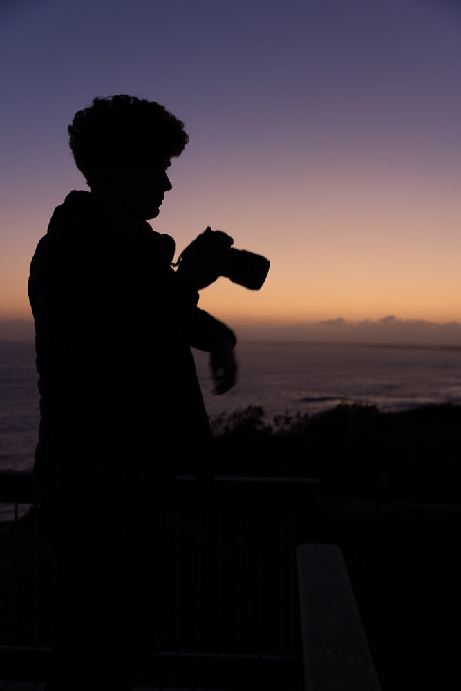 Silhouette of a person with curly hair holding a camera against a twilight sky with the ocean in the background.