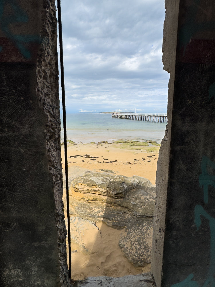 View of a pier extending into the ocean, framed by rough concrete structures with graffiti. The sky is cloudy, and the beach in the foreground has sand and rocks.