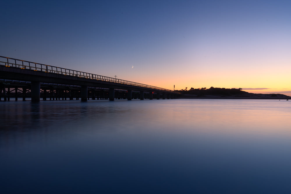 A long bridge stretches across calm water at dusk. The sky transitions from deep blue to a soft orange glow near the horizon, with a sliver of moon visible. The water reflects the colors of the sky and the bridge's lights.