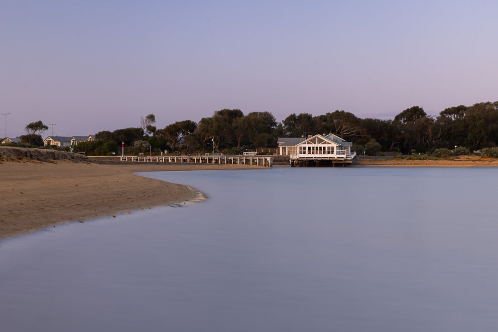 A serene coastal scene unfolds at dusk, with a modern white building on stilts overlooking calm, glassy water. The sandy beach curves into the foreground, leading the eye towards the structure nestled amongst lush trees. The sky above transitions from a soft pink to a pale blue, creating a tranquil atmosphere.