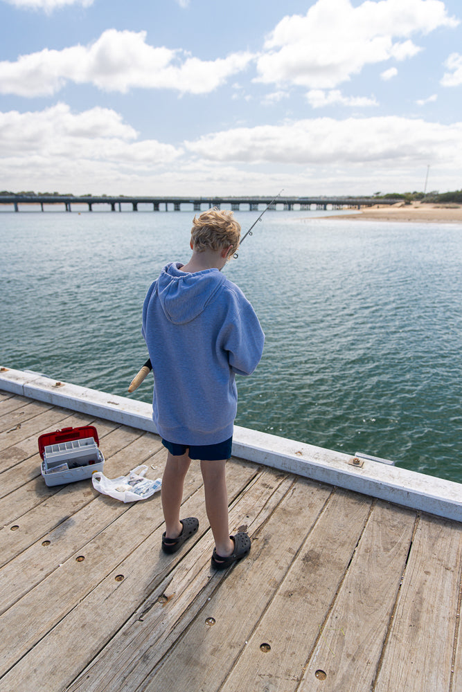 A young boy with blond hair stands on a wooden pier, facing away from the camera, holding a fishing rod. A tackle box and a plastic bag are on the pier beside him. The water is calm, and a long bridge is visible in the distance under a partly cloudy sky.