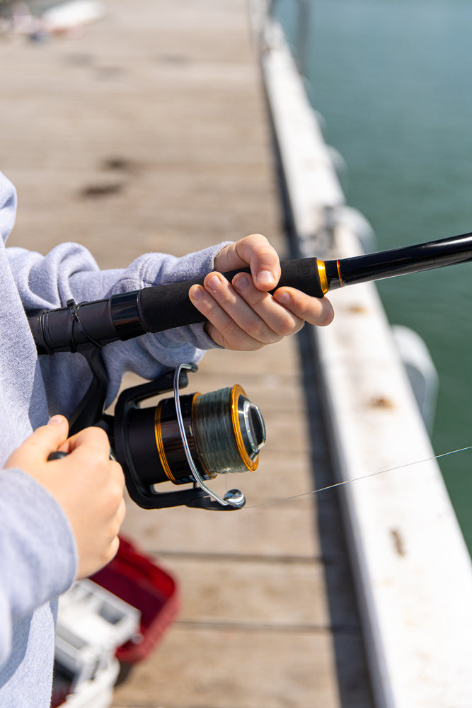 A person wearing a grey hoodie holds a fishing rod with a reel, ready to cast into the water from a wooden pier.