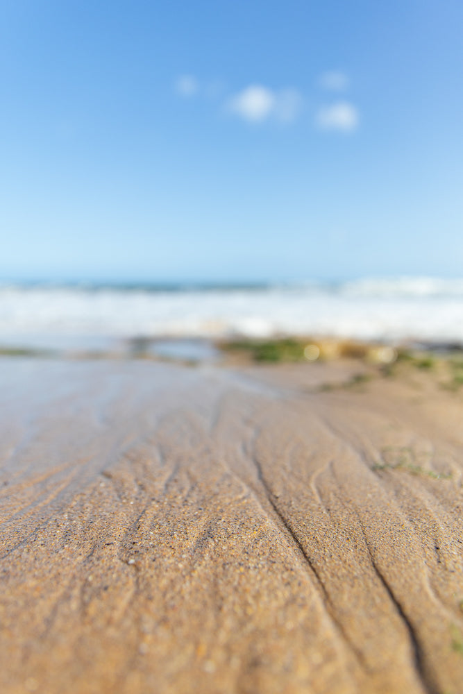 A low-angle shot of wet sand on a beach with ripples from receding water. The ocean and a clear blue sky with a few wispy clouds are visible in the blurred background.