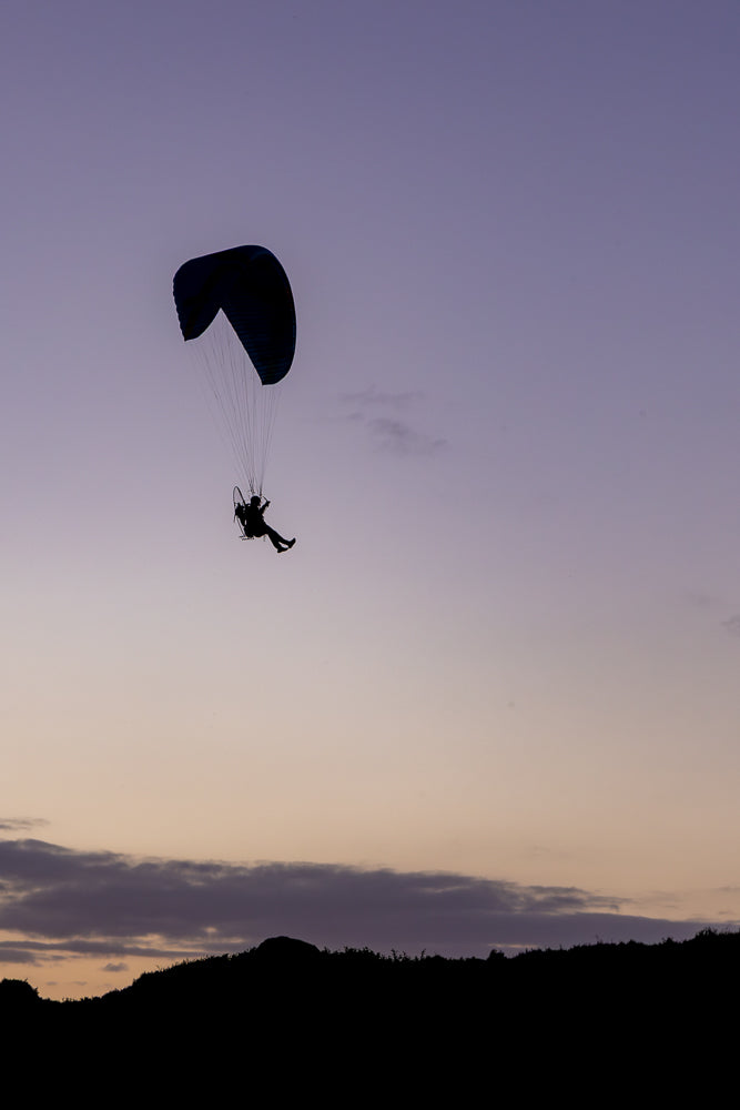 A silhouette of a paraglider in mid-air against a purple and orange twilight sky. The paraglider is suspended beneath a dark blue parachute, with the pilot visible in a harness. The dark silhouette of a hilly landscape with vegetation forms the bottom of the frame.