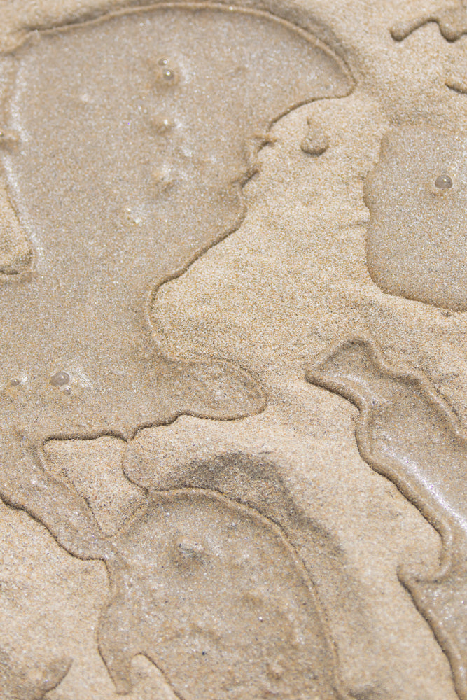 Close-up of wet sand with abstract patterns formed by receding water. The sand is light brown and textured, with shimmering highlights from the moisture. The patterns are organic and flowing, resembling natural contours or ripples.