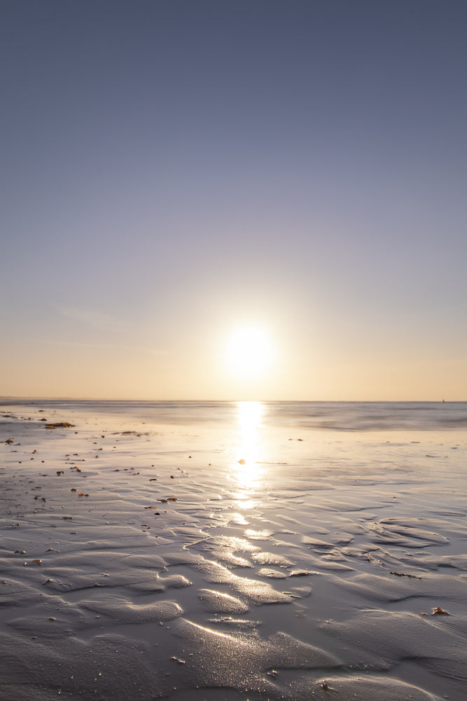 A serene beach scene at sunset. The sun, low on the horizon, casts a golden reflection across the wet sand, creating ripples and patterns. The sky transitions from a pale blue to a warm yellow near the sun.
