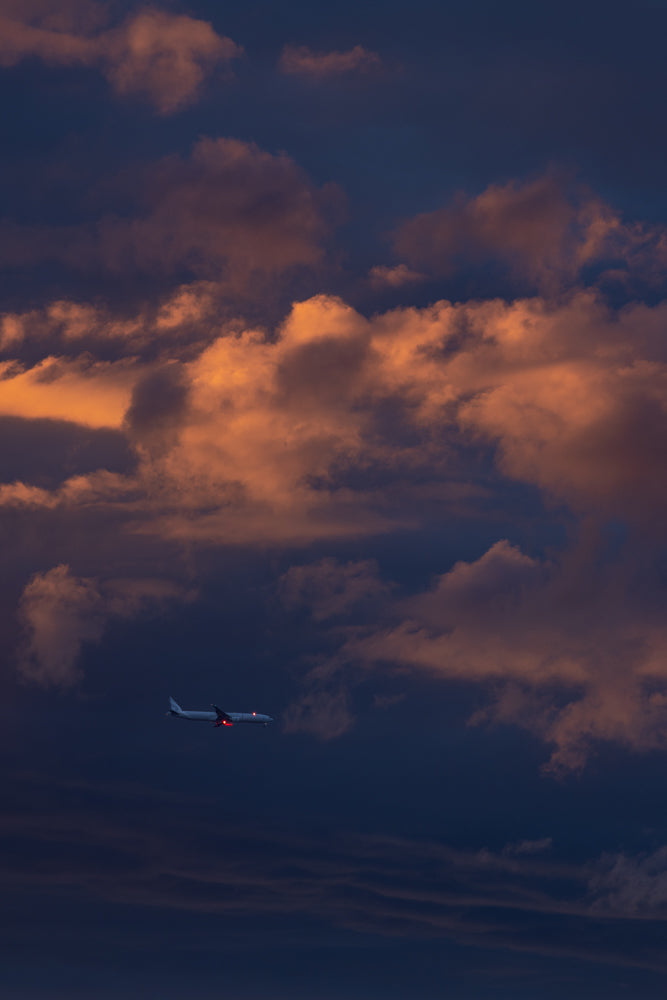 A commercial airplane flies through a dramatic sky filled with clouds illuminated by the warm glow of sunset.