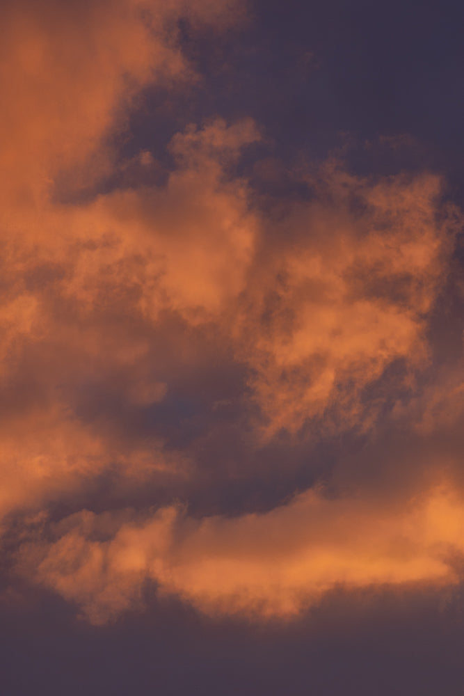 A dramatic sky filled with fluffy, orange-tinted clouds against a deep purple background. The clouds are illuminated from within, creating a warm and vibrant contrast with the darkening sky.