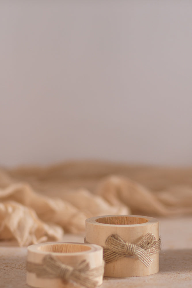 Two wooden napkin rings with twine bows sit on a textured surface, with a soft, draped fabric in the background.