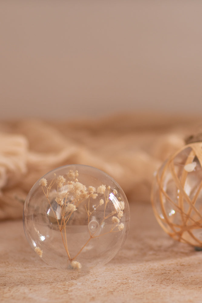 Two neutral organic style glass Christmas ornaments sit on a textured surface. One has baby breath inside, and the other is wrapped in thin, light-colored reeds.